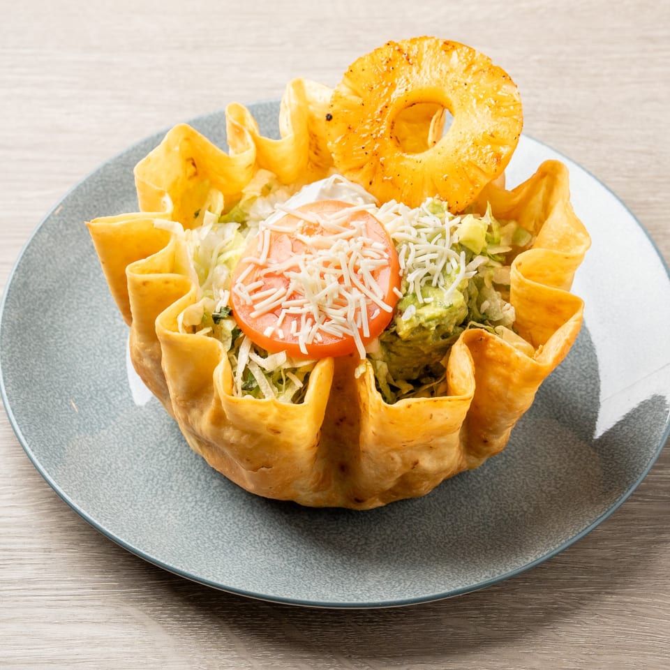 A taco salad in a crispy tortilla bowl, topped with lettuce, tomato, cheese, guacamole, and pineapple.